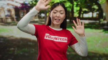 Güneşli bir günde, Çin 'de bir parkta gönüllü olarak çalışan bir kadın kırmızı bir gömlek giyiyor ve yeşil yemyeşil bir zemine karşı neşeyle gülümsüyor, neşe ve coşku ifade ediyor..