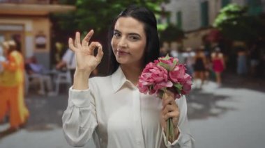 Sokakta elinde canlı bir çiçek buketi olan genç bir İspanyol kadın güzel bir jest yapıyor, şehir ortamında pozitifliği ve zarafeti somutlaştırıyor..