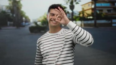 Şehir sokaklarında gülümseyen genç bir adam, çizgili bir gömlek giyiyor, elleriyle iyi bir jest yapıyor, kendine güven ve neşe hissi veriyor..