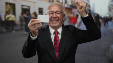 Takım elbiseli üst düzey bir adam, şehir içi iş ortamında başarı ve mutluluğu simgeleyen bulanık yayalarla dolu kalabalık bir sokakta elinde kart tutarak başarıyı kutluyor..