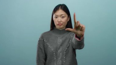 Mavi arka planda ezik jestler yapan kadın, süveter ve el hareketleriyle gençlik ifadesini vurguluyor, kadın ifadesine ve izole ortama odaklanıyor.