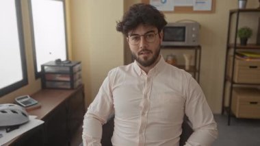 Sakallı genç bir adam modern bir ofiste gözlük takıyor. Etrafı kitaplık, kask ve mikrodalga fırınla çevrili. Rahat, profesyonel bir ev ortamı yaratıyor..