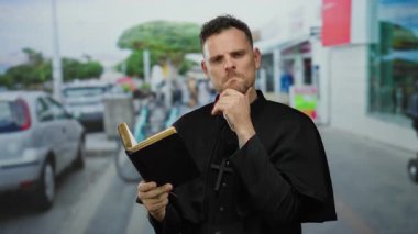 Şehir caddesinde duran bir rahip, siyah bir cübbe giyiyor, elinde bir İncil tutuyor ve şehir bölgesinde derin bir şekilde düşünüyor, inancını ve düşüncelerini dışa vuruyor..