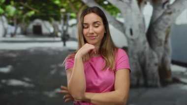 Pembe tişörtlü bir kadın, güneşli bir günde, açık havada, şehir sokağındaki bir ağacın altında neşeli bir ifade sergiliyor..