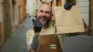 Gülümseyen adam şehir sokaklarında kağıt torba tutuyor. Önlük dövmeleri ve eldivenler takıyor. Bu da modern teslimat servisini işaret ediyor..