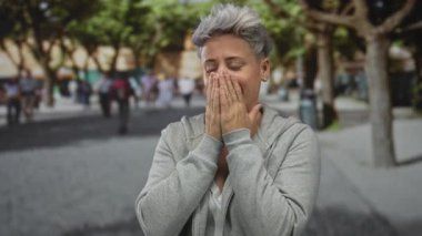 Kadın, gün ışığında bulanık yayaların ortasında şehir caddesinde elleri ve ağzı kapalı bir şekilde ağzını ve burnunu kapatıyor..