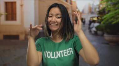 Çinli bir kadın, yeşil tişört giyip, parlak bir şekilde gülümseyen, şehir caddesinde elleri ile çerçeveli jestler yapıyor..