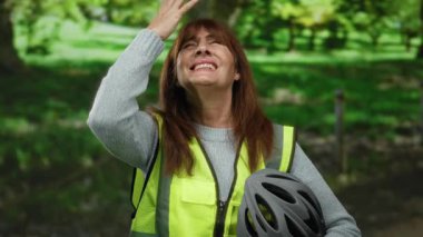 Parktaki son sınıf öğrencisi İspanyol kadın yansıtıcı yelek giyiyor ve elinde bir kask tutuyor. Yeşillik altında duygularını ifade ediyor..