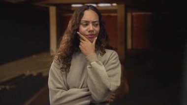 Kapalı bir bina koridoru altında dururken, çene ve kol çapraz el ile genç Afrikalı Amerikalı kadın, gündelik sweatshirt giyen üst ışıklar altında; şüphe üzerine düşünme.