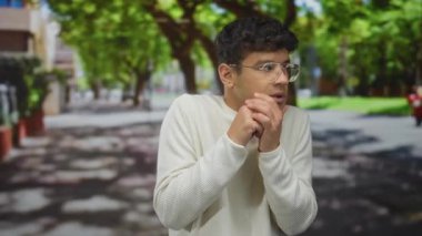 Krem rengi süveter giyen ve yuvarlak gözlükler takan bir adam, yeşil ağaçların altında göğsüne yapışmış eller, baş aşağı dönüyor, endişe özlemi.