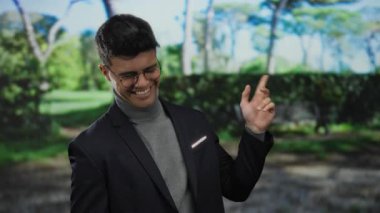 Açık hava parkında gülümseyen İspanyol bir adam gözlüklü ve günlük ceketli, kendine güvenen ve doğal çevrede çekici bir tavır sergiliyor.