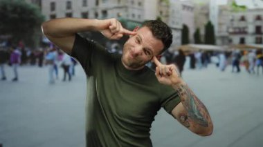 Dövmeli genç bir adam şehir meydanında komik suratlar yapıyor. Arka planda yürüyen insanlar, canlı bir şehir havası ve sıradan bir atmosfer sergiliyor..