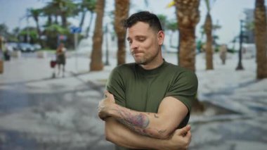 Şehir sokağında palmiye ağaçlarıyla dikilen, yeşil tişörtlü, kolunda dövme olan, düşünceli ve kendini kucaklayan bir adam..