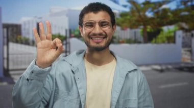 Gözlüklü ve ceketli bir adam sokak girişinde geniş gülümseyerek 