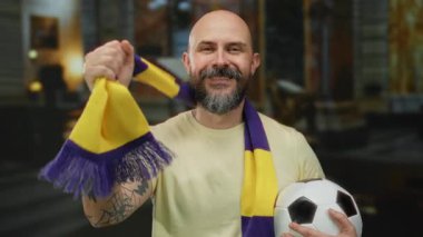 Kapalı alanda gülümseyen sakallı bir adam futbol topu tutuyor ve canlı kilise ortamında renkli bir eşarp takıyor neşeli ve destekleyici bir atmosfer sergiliyor..