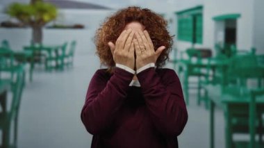 Kıvırcık esmer saçlı İspanyol kadın yeşil mobilyalı bir kahve restoranının terasında gülümsüyor..