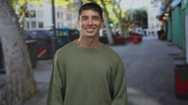 Gevşek yeşil süveter giyen bir adam, şehrin merkezinde ağaçlarla kaplı bir sokakta gülümseyerek kafaya dokunur..