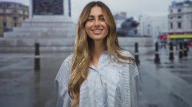 Sarışın kadın çizgili tişört giyer gülümser ve şehir meydanında elini öne uzatır; davetiye açıklığı.