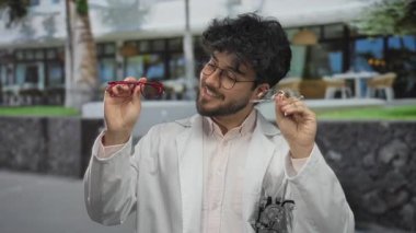 Güneşli bir günde, dışarıda gözlük tutan sakallı genç bir adam. Parlak güneş ışığına hangi çerçevenin daha iyi uyacağını seçerken yüzünde bir endişe ifadesi var.