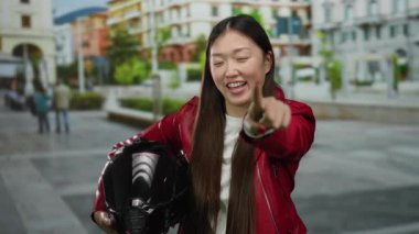 Şehir caddesinde kırmızı ceketli bir kadın motosiklet kaskı tutuyor. Bulanık binalar ve arka planda insanlar var..