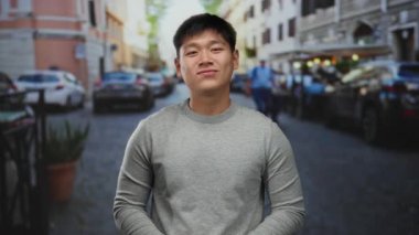 Kalabalık bir şehir caddesinde gülümseyen genç adam, neşeli ve huzurlu bir ruh halini yansıtıyor. Arabalar ve binalar arasında, gri bir süveter giyiyor..