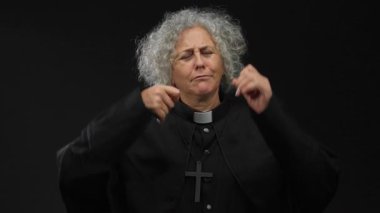 Dini kıyafetleri içinde gri saçlı yaşlı bir kadın siyah arka plana karşı ağlayan bir jest yapıyor. Duygularını ve iç gözlemlerini aktarıyor..