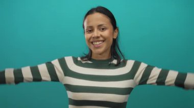 Genç İspanyol kadın gülümsüyor, elleri parlak yeşil bir duvar arkasında kalp şekli oluştururken, çizgili bir gömlek giyiyor ve neşeli görünüyor..