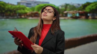 Parkta bir pano tutan gözlüklü bir kadın. Arka planda ağaçlar ve su var. Açık havada profesyonel bir anı yakalıyor..