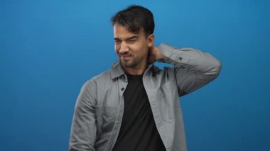 Gri tişörtlü genç İspanyol bir adam rahatsız bir şekilde boynuna dokunuyor mavi izole edilmiş arka planda acı ve huzursuzluk gösteriyor..