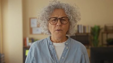 Ofiste kıvırcık gri saçlı ve gözlüklü bir kadın, el hareketleriyle düşünceliliğini ifade eder, kapalı bir arka plan olan kitap ve bitkilerin arasında günlük kıyafetlerini giyer..