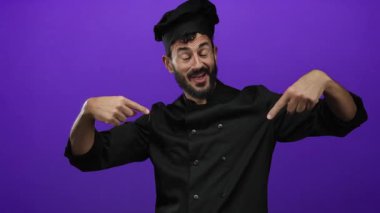 Sakallı İspanyol aşçı, siyah üniformalı ve aşçı şapkalı canlı bir mor arka planda mutfak ortamında kendine güveni ve coşkuyu ifade ederek kendisini işaret ediyor..