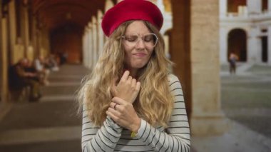 Eski bir atari salonunda bileğini kontrol eden bir kadın uzun koridorda endişeli görünen çizgili bir bluzun üzerine sarı bir bere ve gözlük takıyor..