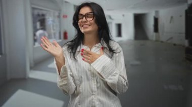 Beyaz çizgili gömlek giyen, el sallayan ve boş stüdyoda gülümseyen bir kadın..
