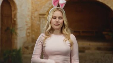 Sarı saçlı, tavşan kulaklı ve pembe bluzlu bir kadın üniversite kampüsünde eski bir tuğla arka planda bir şey gösteriyor..