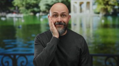 Parkta bir adam yüzüne sürpriz bir ifadeyle dokunuyor, sıradan bir süveter giyiyor, yemyeşil manzaraya ve gölün arka planına karşı duruyor..