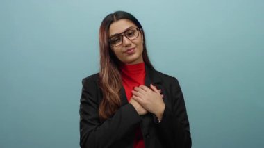 Gözlüklü genç İspanyol bir kadın ve elleri yüreğinde siyah bir ceketle izole edilmiş beyaz bir arka plan duvarına şükranlarını sunuyor..