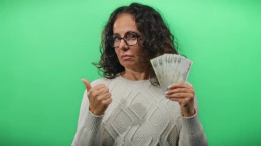 Kafası karışmış bir ifadeyle Dirham banknotları tutan ve sağlam yeşil bir zemine karşı baş parmak kaldıran bir kadın belirsizlik ve mali karar alma duyusunu iletiyor..