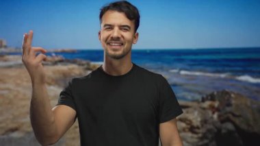 Genç İspanyol bir adam açık mavi bir gökyüzünün altında arka planda okyanusla deniz kıyısında gülümsüyor.