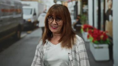 Hispanic senior woman wearing glasses gestures okay sign on a bustling city street with blurred background of red flowers and vehicles, embodying confidence and positivity.