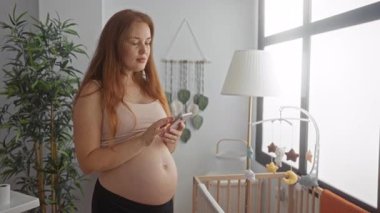 Hamile kadın çıplak karnını okşar ve parlak güneşli kreş odasında ahşap beşik ile akıllı telefon ekranına dokunur..