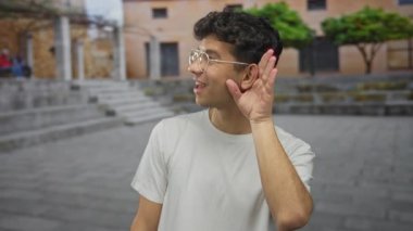 Beyaz tişörtlü ve gözlüklü bir adam taştan bir sokakta kulağını kavrıyor..