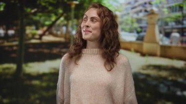 Gülümseyen ve el kol hareketi yapan genç bir kadın açık hava parkında yemyeşil ağaçlar ve şehir manzarasıyla neşeli ve rahat bir atmosfer yakalıyor..