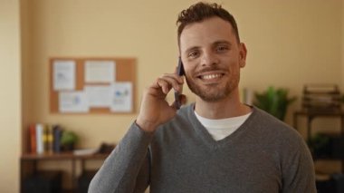 Ofiste telefonla konuşurken gülümseyen bir adam, arka planda bir ilan tahtası ve bitkileri olan profesyonel bir iç mekan ortamına güvenerek..