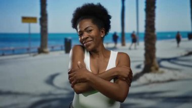 Afrika kökenli Amerikalı bir kadın, arka planda palmiye ağaçları ve okyanusla güneşli bir sahil yürüyüşünde sıcaklığını ve kendini sevgini ifade ediyor..
