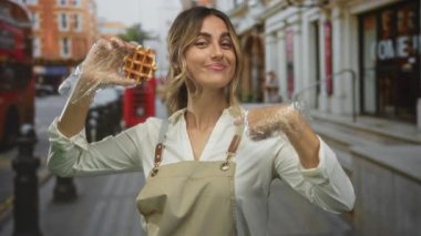 Sarışın genç kadın, eldivenli elleriyle waffle tutuyor ve şehir caddesinde waffle yapmak için işaret ediyor; davetiye keyfi.