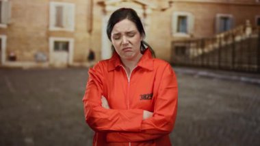 Turuncu hapishane tulumlu esmer kadın üniversite kampüsünde kollarını kavuşturarak duruyor ve tarihi tuğla mimarisinin verdiği hayal kırıklığını dile getiriyor..