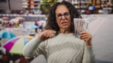 Orta yaşlı bir kadın, renkli şemsiyeler ve bulanık insanlarla dolu kalabalık bir plaj ortamında, hayal kırıklığına uğramış bir ifadeyle kulağını kapatarak deniz kenarında Mısır banknotları tutuyor..