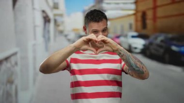 Genç adam şehir caddesinde el kalp hareketi yapıyor, çizgili tişört giyiyor, mutlu bir şekilde gülümsüyor, dövmeler görünür, rahat bir şehir havası yakalıyor..
