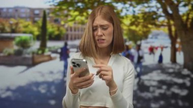Sarışın genç bir kadın, güneşli bir caddede ağaçlı ve arka planda bulanık insanlar olan karışık bir yüz ifadesiyle akıllı telefon kullanıyor..