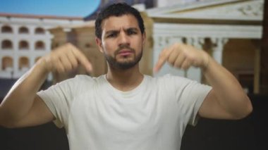 Beyaz tişörtlü adam mavi stüdyoda işaret parmaklarını sıkıca aşağıya doğru yönlendiriyor..